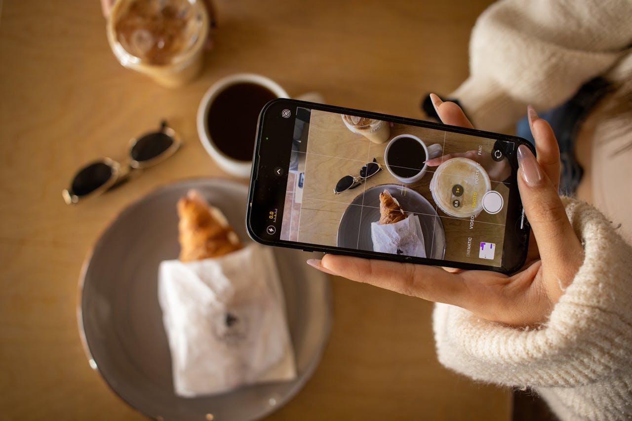 services-01 Woman photographing coffee and croissant with smartphone at a cafe.
